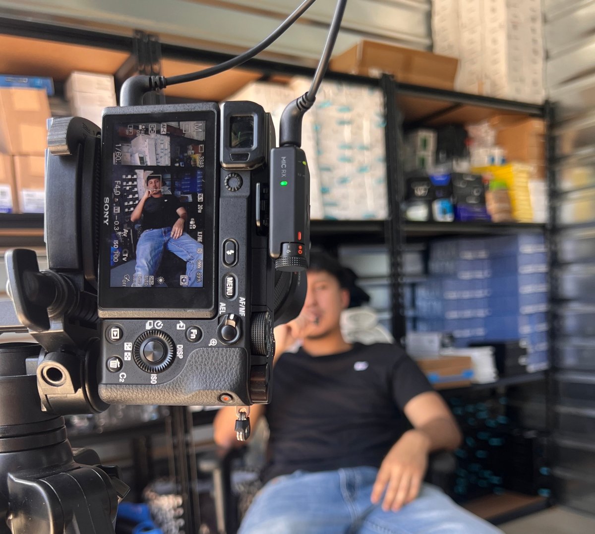 Professional cinema camera on tripod displaying live feed of a man sitting in a studio with shelving in background