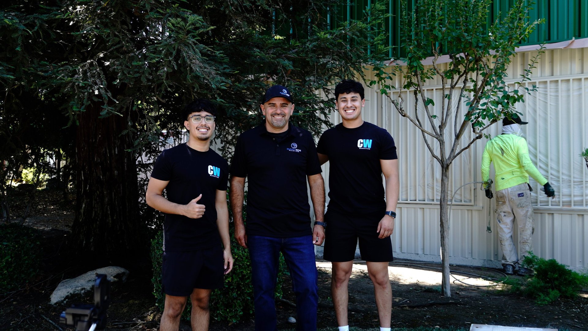 Three people wearing matching black polo shirts stand together outdoors in front of a white metal gate and dense green foliage