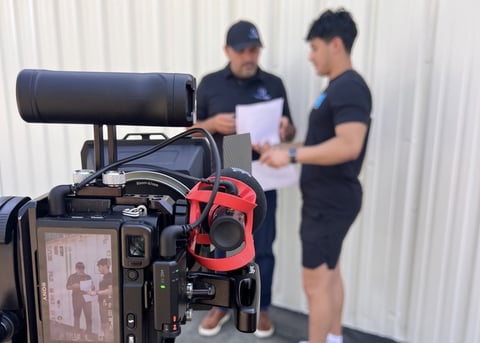 Professional video camera setup with microphone in focus, two crew members in black shirts collaborating in background